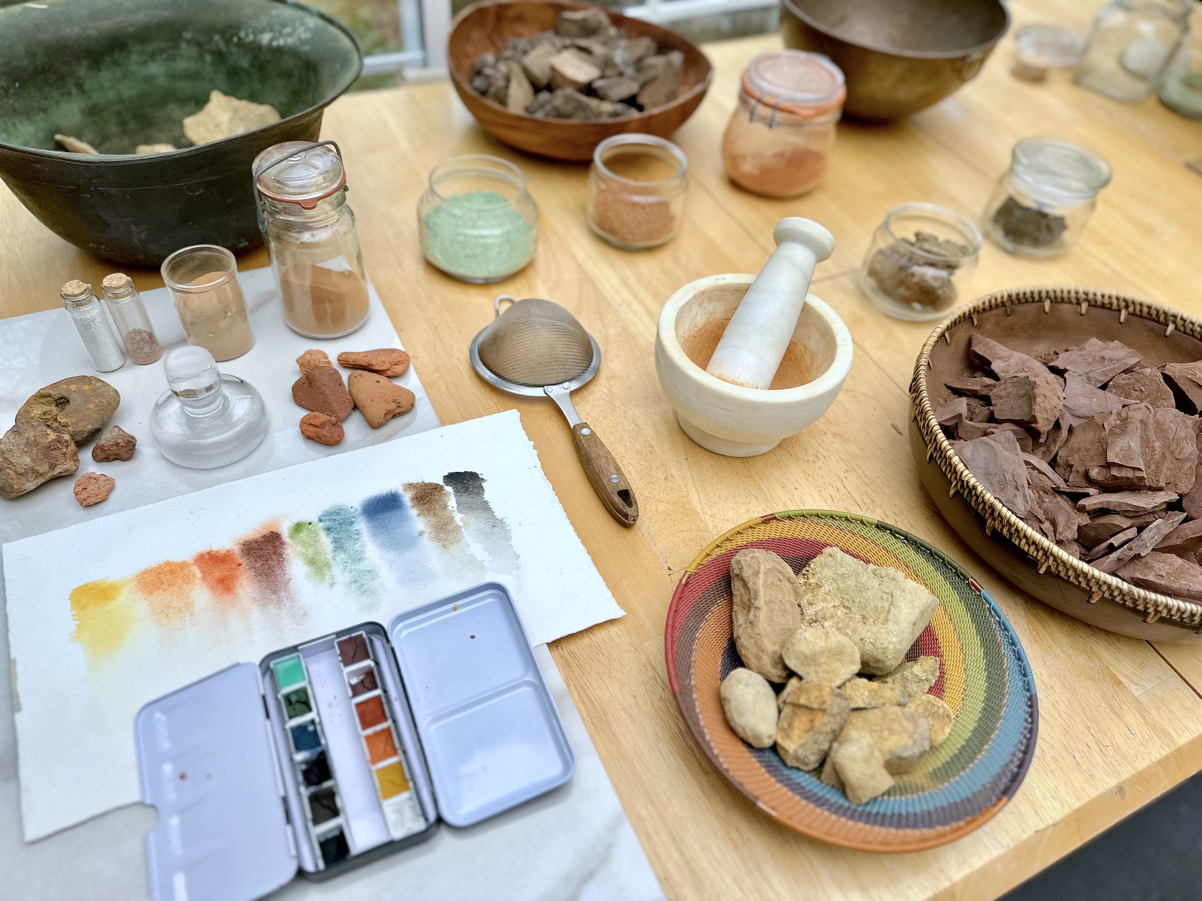 Natural pigment‑making materials arranged on a table, including rocks, powders, jars, tools, and color swatches.