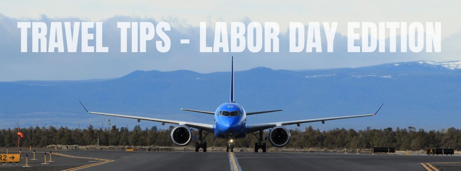 A bright blue commercial airplane is centered on a runway, facing forward with its landing gear down. The plane sits against a backdrop of distant blue mountains and a cloudy sky. Large white text across the top reads “TRAVEL TIPS – LABOR DAY EDITION.” Yellow runway signs and markers are visible along the pavement in the foreground.
