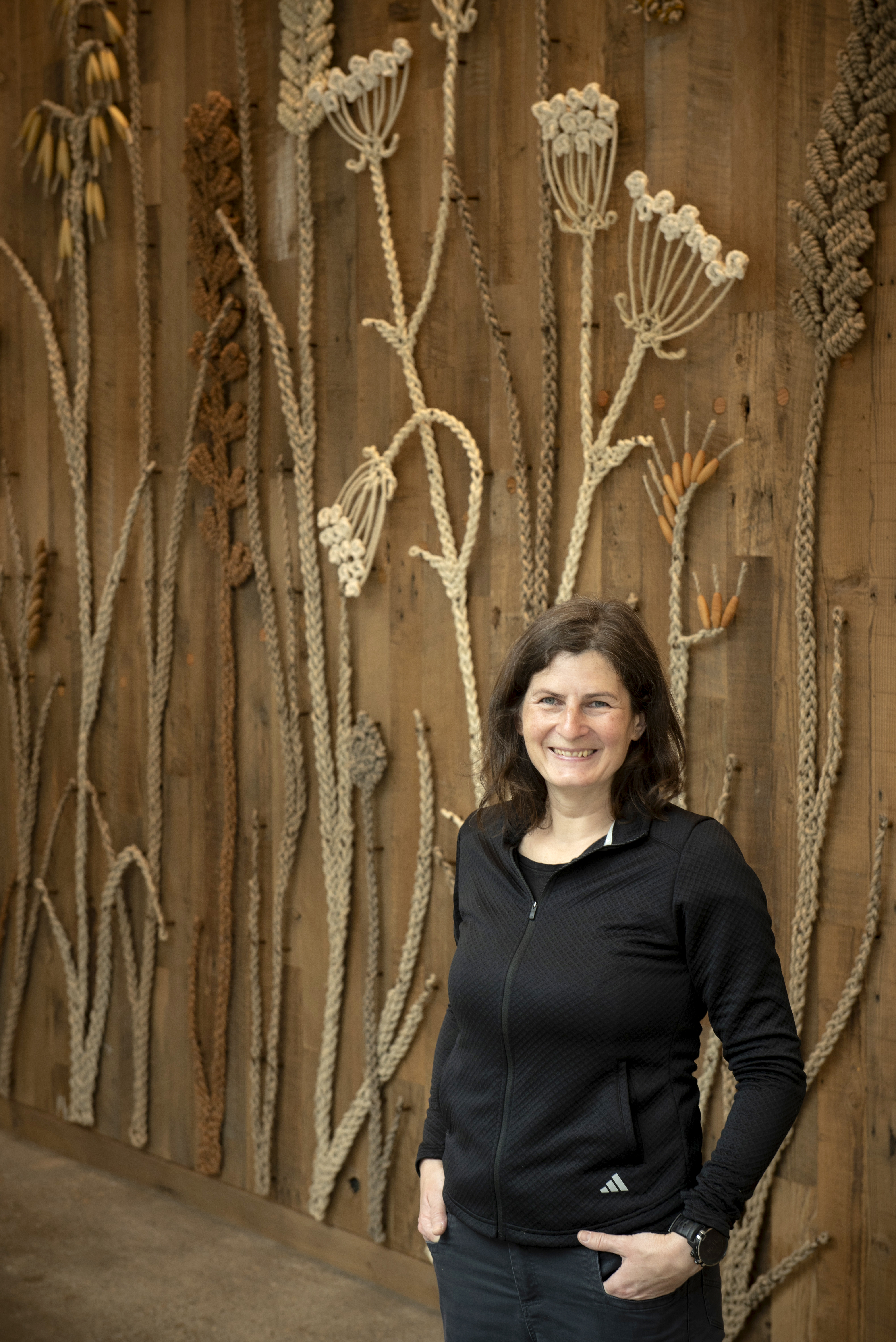 Artist Manca Ahlin standing in front of a large wall installation made of woven natural fibers shaped into botanical forms on a wooden backdrop.